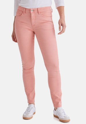 Frau trägt eng anliegende pinke Jeans und weiße Turnschuhe, steht vor weißem Hintergrund.