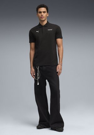 Man staat en draagt een zwart Puma McLaren poloshirt, zwarte wijd uitlopende jeans met witte oordopjes die uit de zak hangen, en zwarte schoenen.