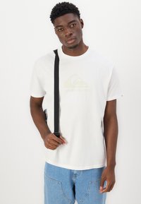 Hvit bomulls T-skjorte med et lysfarget logo, kombinert med blå denim jeans og en svart skulderveske. Enkelt moderne design.