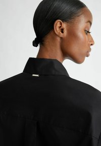 Camisa negra con un cuello estructurado, que presenta una pequeña etiqueta metálica en la parte posterior. La tela parece suave y de textura mate.