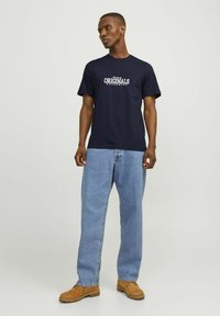 Camiseta de algodón azul marino con un gráfico blanco de "ORIGINALS". Combinada con unos vaqueros sueltos y botas marrones.