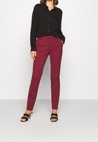 Chemise noire à manches longues avec boutons, associée à un pantalon ajusté bourgogne et des sandales noires à talons avec un design tressé.