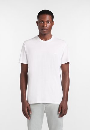 Mladý muž stojaci vpred, oblečený v jednoduchom bielom tričku s krátkymi rukávmi a svetlosivých nohaviciach na bielom pozadí.