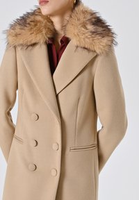 Cappotto beige doppiopetto in tessuto testurizzato con un grande colletto in pelliccia nei toni del tan e del marrone. Presenta due tasche frontali e bottoni.