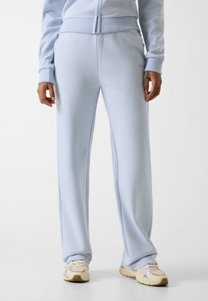 Osoba ubrana w jasnoniebieskie, luźne spodnie dresowe i pasującą kurtkę, oraz beżowo-białe sneakersy, stojąca na jednolitym tle.