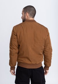 Bruine nylon bomberjack met geribde halslijn en manchetten, voorzien van zijzakken en een gladde, matte textuur. Draagt zwarte broek.