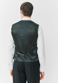 Fekete selyemmel bevont mellény sima textúrával, keskeny hátrészülettel és testre szabott szabással, fehér, hosszú ujjú ing felett.