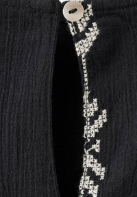 Pleteni črn kardigan z teksturirano tkanino, sprednjim zapiranjem na gumbe in belimi vezenimi cvetličnimi poudarki ob odprtini.