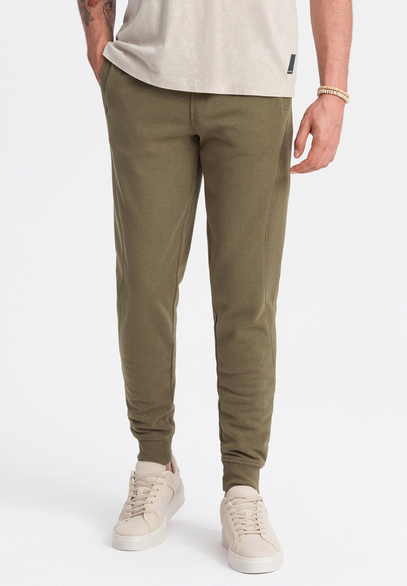 Olijfgroene sweatpants van zacht materiaal, met elastische manchetten en zijzakken, gecombineerd met lichtbeige sneakers.