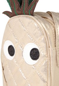 Zaino trapuntato dorato con grandi occhi bianchi, pupille nere scintillanti e foglie di filato verde che somigliano a un ananas. Dettagli della chiusura con zip visibili.