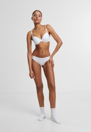 Balts mīkstināts krūšturis un saskaņotas bikini biksītes no gluda auduma. Modelis stāv ar vienu roku uz gurniem, valkājot baltas potīšu zeķes ar viļņotām malām.