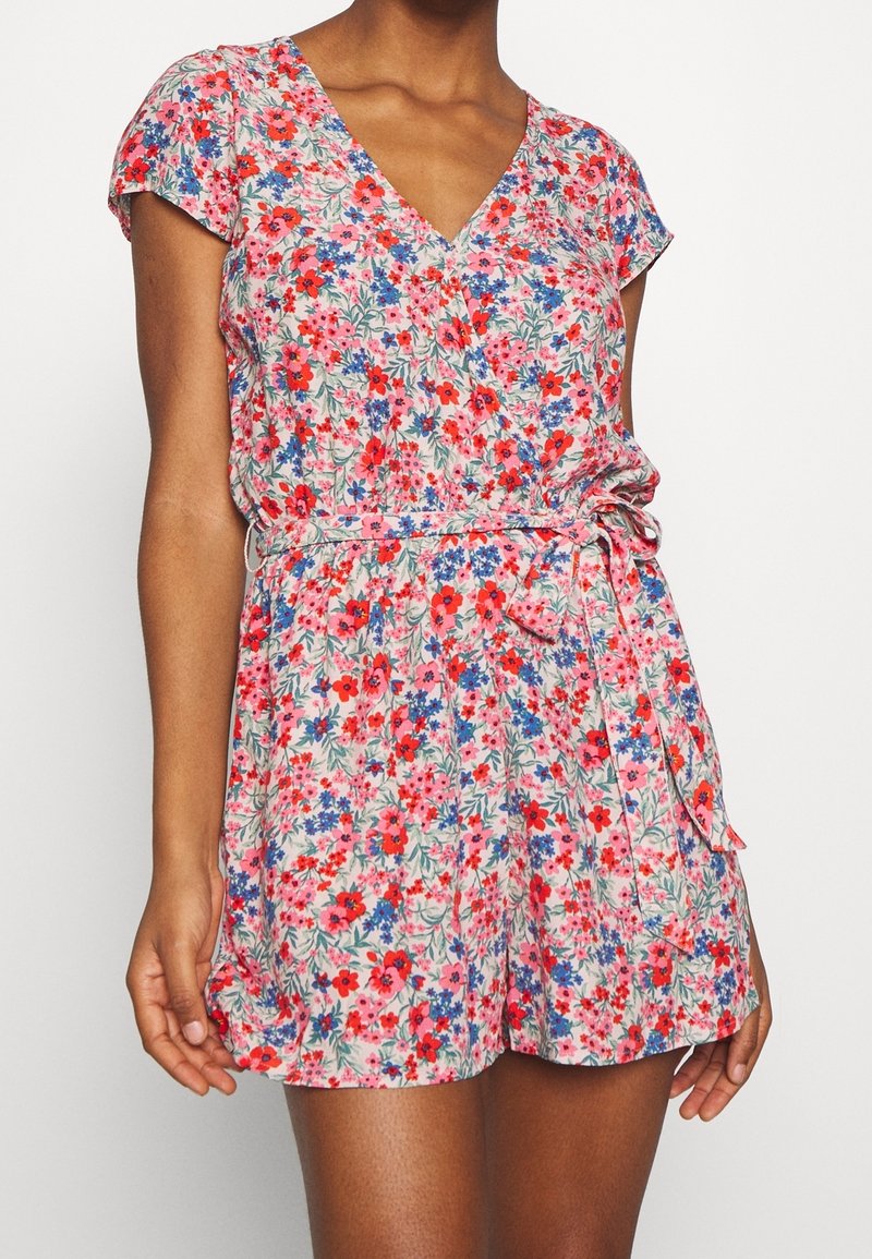 Femme portant une combinaison courte à manches courtes fleurie avec des fleurs rouges, roses et bleues, col en V et ceinture nouée à la taille.