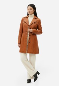 Brun läder trenchcoat med krage, två knappar och sidofickor, bärs över en krämfärgad polotröja och ljusa byxor, kombinerat med svarta stövlar.