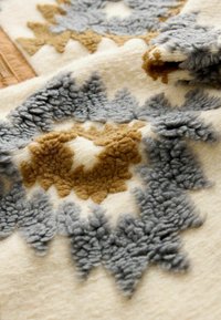 Wolltextil mit cremefarbenem Grund und grauen sowie braunen, texturierten abstrakten Mustern. Die Oberfläche ist plüschig und weich mit erhöhten Fasern.