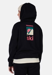 Felpa nera con grafica sul retro che presenta montagne e un sole in rosa e verde mare. Maniche e orlo a coste. Indossata con una maglietta chiara sotto.
