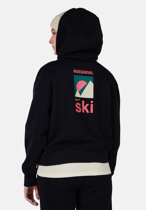 Zwarte hoodie met een afbeelding op de achterkant van bergen en een zon in roze en teal. Geribbelde manchetten en zoom. Draagt een lichtgekleurde onderhemd.