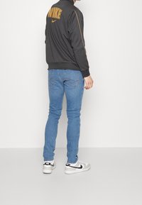 Person som bär en svart Nike-jacka med orange logotyp, ljusblå jeans och vita Nike-sneakers stående mot en helt vit bakgrund.