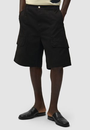 Uomo che indossa pantaloncini cargo neri fino al ginocchio con grandi tasche laterali e scarpe di pelle nere senza lacci, su uno sfondo semplice.