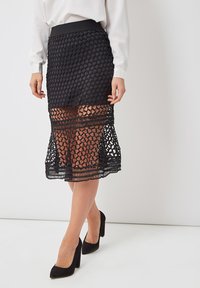 Falda de encaje negro con patrón geométrico, con cintura ajustada y dobladillo en capas. Combinada con tacones altos negros.