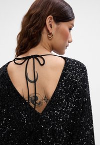 Juodas blizgus vakarinis suknelė su V formos nugaros dizainu, virš nugaros užrišama juoda juostelė. Matoma tatuiruotė ant nugaros, įskaitant mėnulio formas.