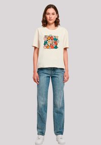Cream cotton t-shirt with a colorful abstract graphic design featuring flowers and smiling faces, paired with light blue jeans and white sneakers.
