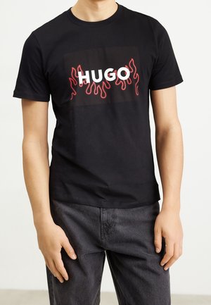 Homme portant un t-shirt noir avec le texte "HUGO" et des graphiques de flammes rouges, associé à un jean gris foncé, debout sur un fond uni.