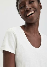 T-shirt bianco a maniche corte con scollo a V profondo. Il tessuto sembra morbido e leggero, con una finitura leggermente strutturata.