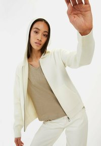 Witte zip-up hoodie met een capuchon en voorzakken, gecombineerd met een beige V-hals top, gedragen met lichte broek, wat zorgt voor een ontspannen pasvorm.
