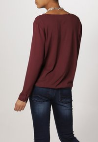 Blusa de manga larga burdeos con escote redondo, fabricada en tejido suave y de corte relajado, combinada con jeans de denim azul oscuro que presentan un patrón de costura simple.
