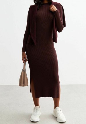 Femme portant une longue robe en tricot côtelé marron avec des fentes latérales, des baskets blanches, un pull noué sur les épaules et tenant un sac à main beige.