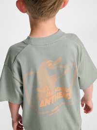 Barn med kort blondt hår iført en grøn t-shirt med en orange tegneseriefugl og tekst på ryggen.