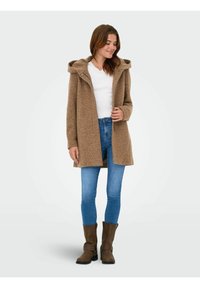 Bruine gestructureerde jas met een capuchon, rits aan de voorkant en zijzakken, gecombineerd met een blauwe jeans en bruine enkellaarsjes.