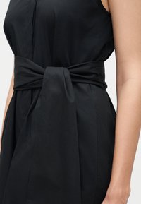 Αμάνικο μαύρο φόρεμα με φαρδιά υφασμάτινη ζώνη δεμένη κόμπο στη μέση σε κορμό ενός ατόμου μπροστά σε απλό φόντο.