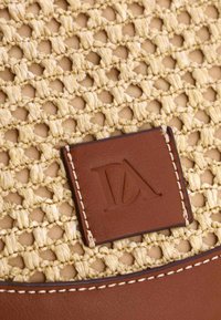 Bolso de paja trenzada con un cuerpo texturizado en tono marrón claro, detalles en cuero marrón oscuro y un parche rectangular con logotipo en relieve.