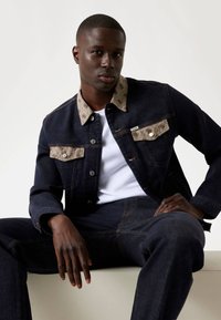 Chaqueta de mezclilla con cuerpo azul oscuro y cuello de color beige con estampado. Cuenta con dos bolsillos frontales y botones plateados. Combinada con jeans oscuros a juego.