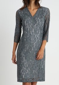 Une robe en dentelle grise jusqu'aux genoux avec des manches trois-quarts, un décolleté en V et des motifs floraux. Le tissu est transparent avec une finition texturée.