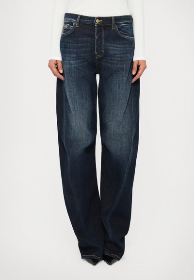 Jean large bleu foncé avec des détails délavés, porté avec un haut à manches longues blanc et des chaussures noires à bout pointu.