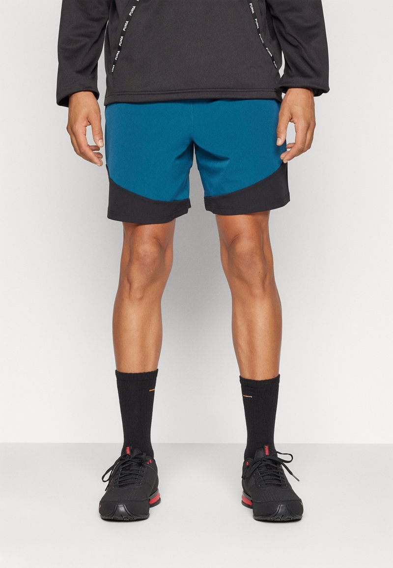 Homme portant un short de sport noir et bleu, un haut à manches longues noir, des chaussettes noires et des baskets noires, debout sur un fond uni.