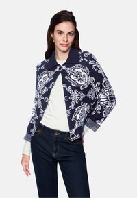 Navy blue cropped cardigan with white paisley pattern, featuring a solid collar and button closure. Worn over a white top and dark jeans.