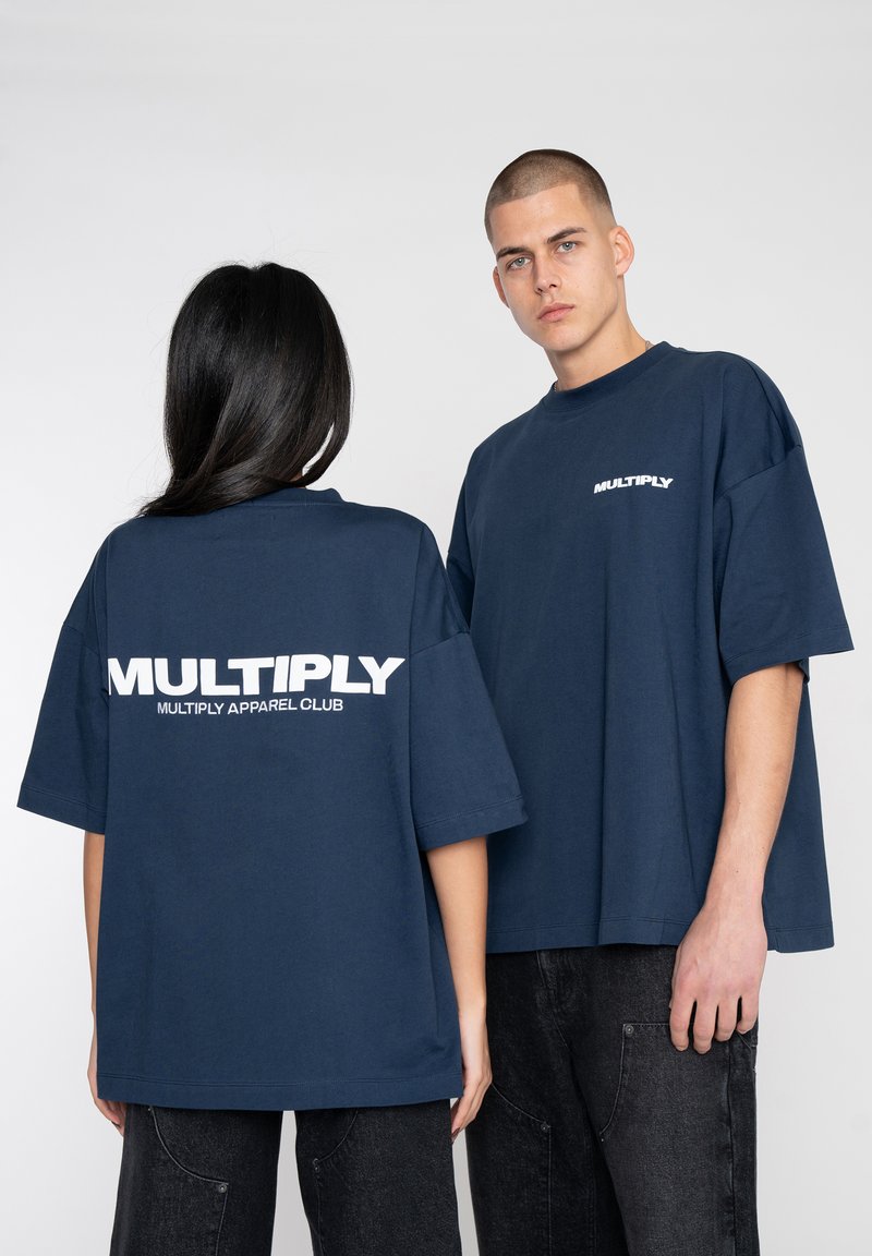 Pimeä sininen ylisuuri t-paita, puuvillamateriaalia, lyhyet hihat, jossa on valkoinen "MULTIPLY" -logo selässä ja edessä, yhdistettynä tummiin housuihin.