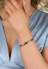 Guldfärgad metallarmband på ett handled, slät yta, cirkulär form, bärs med ett ljusblått satinplagg.