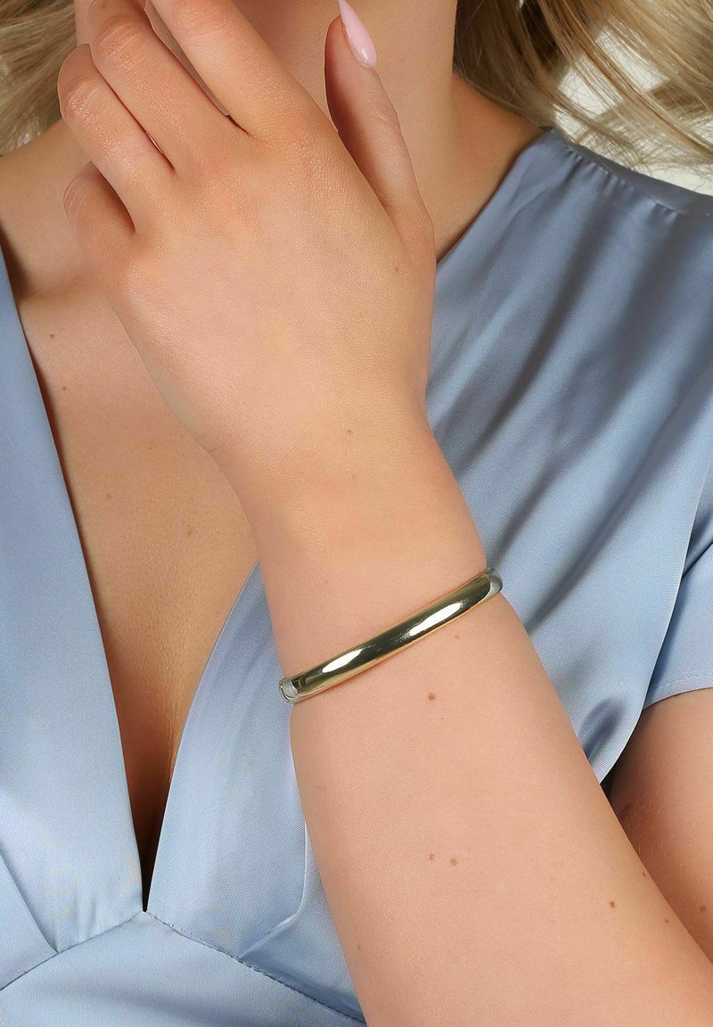 Guldfärgad metallarmband på ett handled, slät yta, cirkulär form, bärs med ett ljusblått satinplagg.