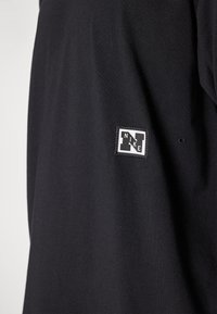 Fekete Nike póló, amelyen egy hímzett fehér címke található a márkanevével és emblémájával. Az anyag simának tűnik, könnyű textúrával.