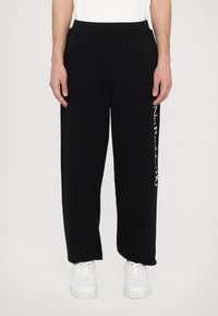 Black sweatpants with a relaxed fit, elastic waistband, and white text on the side. Worn with white sneakers. Smooth texture.