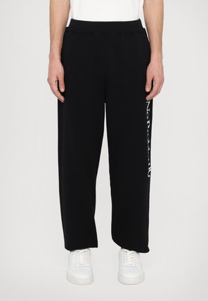 Black sweatpants with a relaxed fit, elastic waistband, and white text on the side. Worn with white sneakers. Smooth texture.