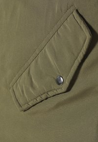 Olivově zelená látka s texturovaným povrchem má šikmou klopovou kapsu zajištěnou kovovým patentem. Viditelné jemné švy.