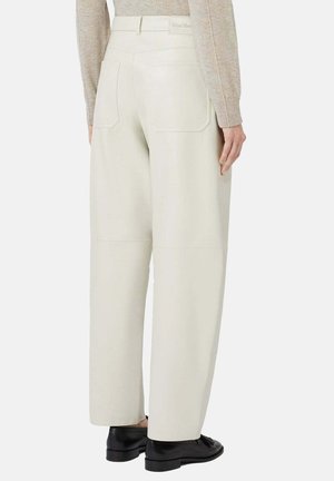 Donna che indossa pantaloni crema a gamba larga e vita alta con tasche posteriori, abbinati a un maglione beige e mocassini in pelle neri, in piedi su sfondo bianco.
