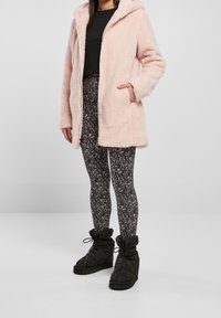 Abrigo con capucha de color rosa claro y esponjoso, con bolsillos frontales. Leggings negros con un patrón abstracto en blanco. Botines negros con un diseño acampanado.