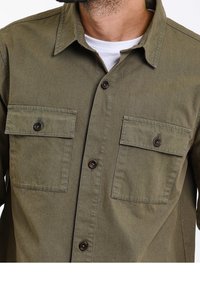 Camisa abotonada verde oliva con dos bolsillos en el pecho, botones marrones y un cuello estructurado. Fabricada con tejido entrelazado de textura suave.