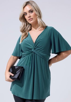 Blondynka w turkusowej sukience z detalem skręconego węzła trzyma czarną kopertówkę, pozując na tle jasnoszarego tła.